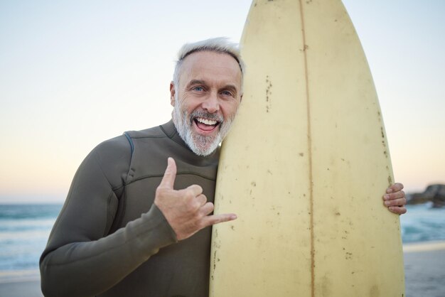 Älterer Mann mit Surfbrett und am Strand lächeln glücklich und bei Sonnenuntergang mit Badebekleidung im Urlaub und Urlaub Portrait Shaka und älterer männlicher Surfer Handzeichen und Surfen am Meer während einer Reise