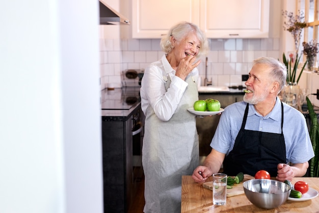 Älterer Mann schnitzt Gemüse und Frau hält Teller mit Äpfeln, kocht zusammen, genießt es, gesund zu sein. Zuhause