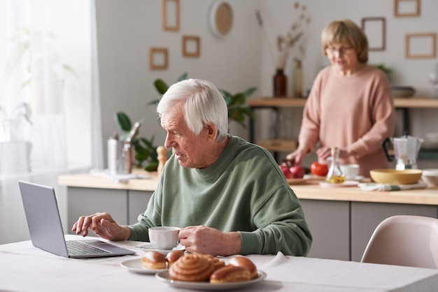 Älterer Mann sitzt am Tisch und tippt auf dem Laptop, der zu Hause online arbeitet, während seine Frau im Hintergrund kocht