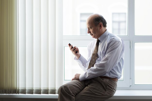 Älterer Mann sitzt auf einer Fensterbank und liest Nachrichten auf Ihrem Smartphone Ruhen Sie sich mitten am Arbeitstag aus