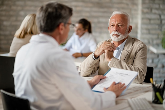 Älterer Mann und Mitarbeiter des Gesundheitswesens kommunizieren während eines Treffens in der Klinik über Krankenversicherungsdokumente