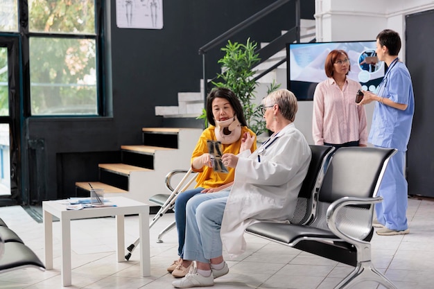 Älterer mediziner und patient mit zervikalem halskragen, der sich eine röntgenuntersuchung ansieht und einen untersuchungsbesuch im wartezimmer des krankenhauses durchführt. Asiatische Frau mit körperlichen Schmerzen und Verletzungen mit Arzttermin.