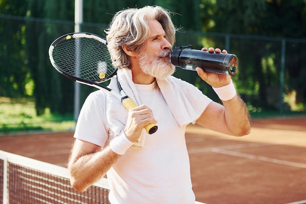 Älterer, moderner, stilvoller Mann mit Schläger im Freien auf dem Tennisplatz bei Tagestrinkwasser