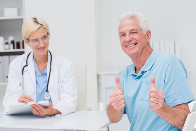 Älterer Patient, der oben Daumen durch Doktor in der Klinik gestikuliert