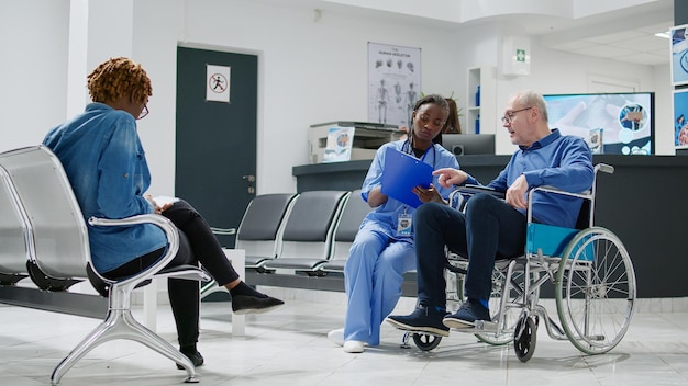 Älterer Patient im Rollstuhl, der sich mit der Krankenschwester in der Lobby des Wartebereichs berät und einen Mann mit chronischer Beeinträchtigung und Behinderung untersucht. Spezialist und Person beim Kontrollbesuch. Stativaufnahme.