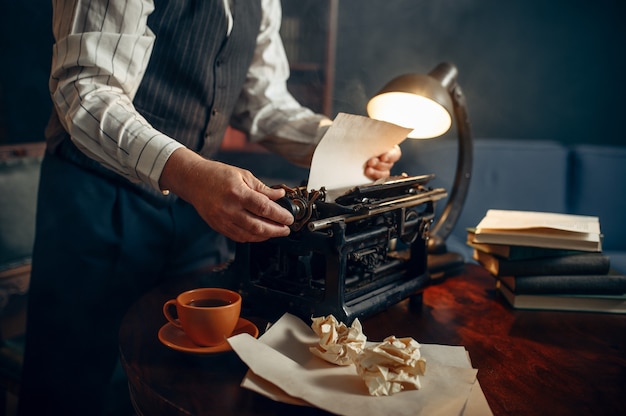 Älterer Schriftsteller fügt Papier in die Vintage-Schreibmaschine in seinem Heimbüro ein. Alter Mann schreibt Literaturroman im Zimmer mit Rauch, Inspiration, Kaffee und zerknitterten Laken auf dem Tisch