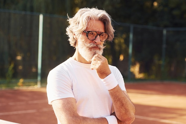 Älterer stilvoller Mann in Brillen, weißem Hemd und schwarzen sportlichen Shorts auf dem Tennisplatz