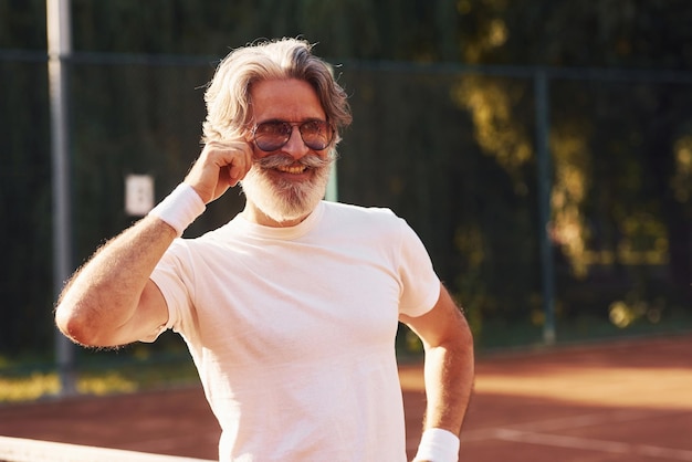 Älterer stilvoller Mann in Brillen, weißem Hemd und schwarzen sportlichen Shorts auf dem Tennisplatz