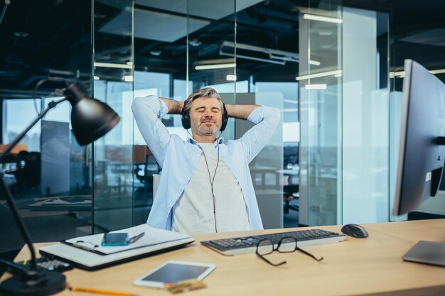 Älterer und erfahrener Geschäftsmann, der sich während einer Pause bei der Arbeit ausruht und Musik vom Kopfhörermanager hört, der in einem modernen Büro arbeitet