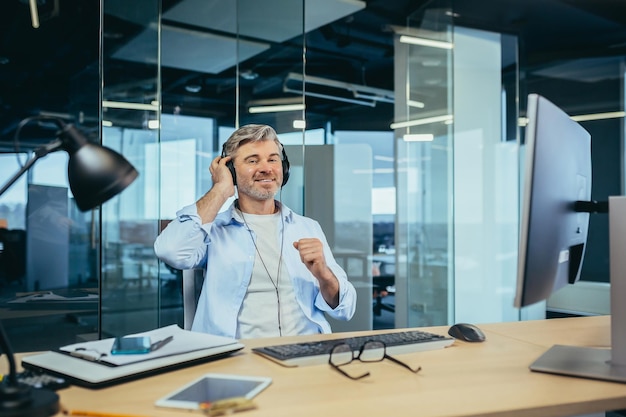 Älterer und erfahrener Geschäftsmann, der sich während einer Pause bei der Arbeit ausruht und Musik vom Kopfhörermanager hört, der in einem modernen Büro arbeitet