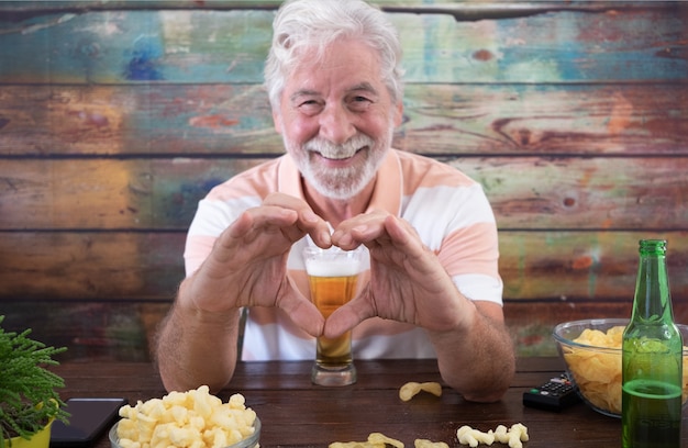 Älterer weißhaariger Mann, der mit Bier und Pommes am Holztisch sitzt, macht Herzform mit den Händen
