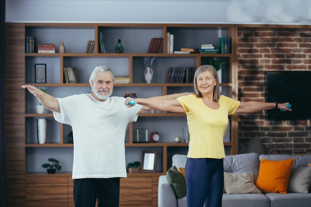 Älteres Ehepaar im Ruhestand, das zu Hause Fitness macht, im Wohnzimmer mit Hanteln steht und in die Kamera lächelt