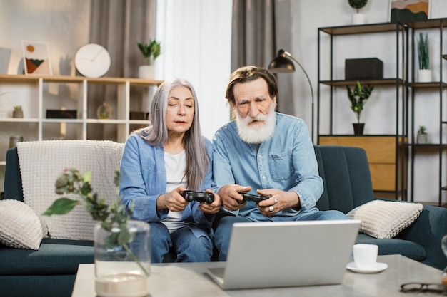 Älteres Ehepaar mit Joysticks in den Händen, das Videospiele spielt, während es auf einer bequemen Couch sitzt