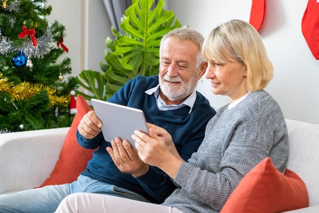 Älteres Ehepaar nutzt Tablet-to-Video-Telefonanruf, um seine Familie zum Weihnachtsfest auf dem Sofa mit Dekoration und Baum zu begrüßen