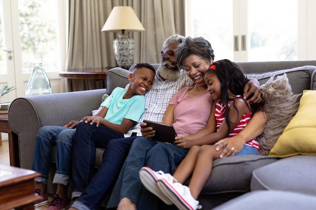 Älteres gemischtrassiges Paar genießt mit seinen Enkelkindern die gemeinsame Zeit zu Hause, sitzt auf einer Couch, umarmt sich und nutzt ein digitales Tablet