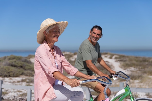 Älteres kaukasisches Paar genießt die Zeit am Strand an einem sonnigen Tag und fährt Fahrrad mit Sand im Hintergrund