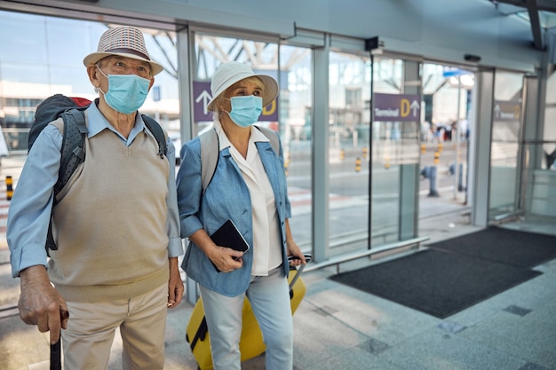 Älteres kaukasisches Paar in Hüten und Einweg-Gesichtsmasken, das zusammen am Flughafenterminal spazieren geht