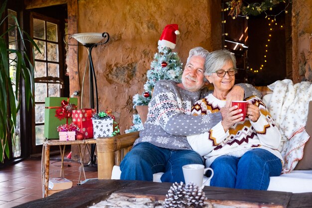 Älteres lächelndes Paar, das zu Hause im rustikalen Chalet zur Weihnachtszeit sitzt und sich auf dem Sofa entspannt. Weihnachtsbaum, Geschenke und Dekorationen im Hintergrund