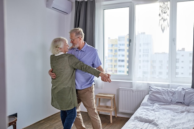 Älteres Paar, das im Wohnzimmer tanzt, Ehemann, der Hand der reifen Frau hält, die Spaß zusammen genießt, Ferien verbringt, Freizeitruhe ifestyle zu Hause. in lässiger Haushaltskleidung