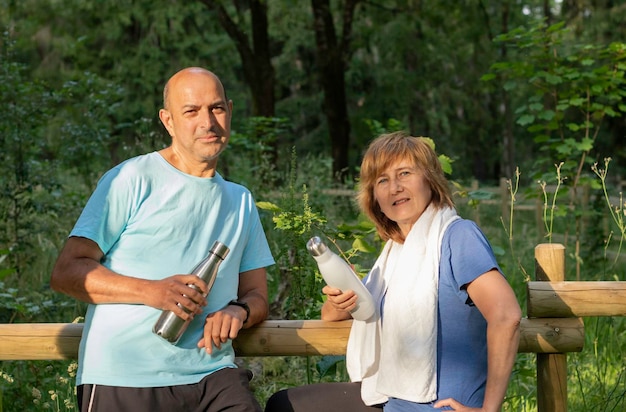 Älteres Paar, das sich nach der Übung im Park mit wiederverwendbaren Metallflaschen in die Kamera lehnt