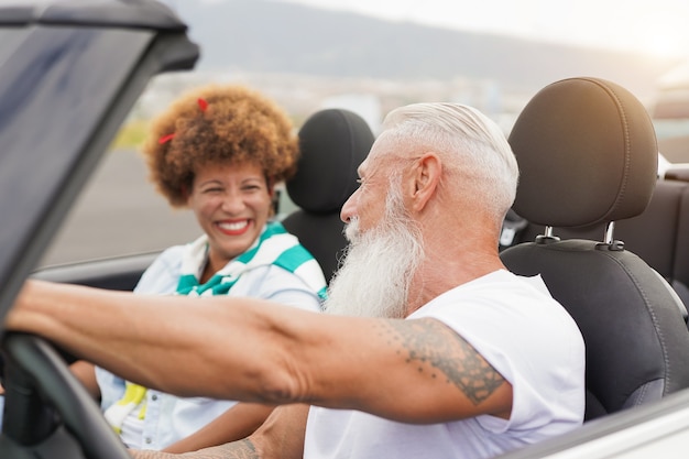 Älteres Paar, das Spaß im Cabrio-Auto hat - Multirassische Senioren auf einem Roadtrip mit Cabriolet-Auto - Sommerferienkonzept