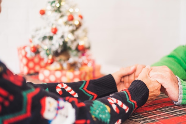 Älteres Paar in warmer Kleidung, das sich zu Hause die Hände vor geschmücktem Weihnachtsbaum hält. Liebevolles altes romantisches heterosexuelles Paar, das zusammen Weihnachtsfest feiert