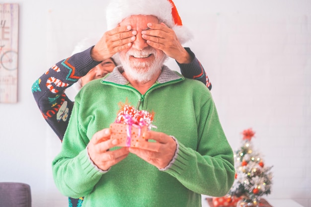 Älteres Paar in warmer Kleidung und Weihnachtsmütze Frau, die die Augen des alten Mannes mit einem Überraschungsgeschenk für ihn bedeckt Liebendes altes romantisches heterosexuelles Paar, das zusammen das Weihnachtsfest feiert xA