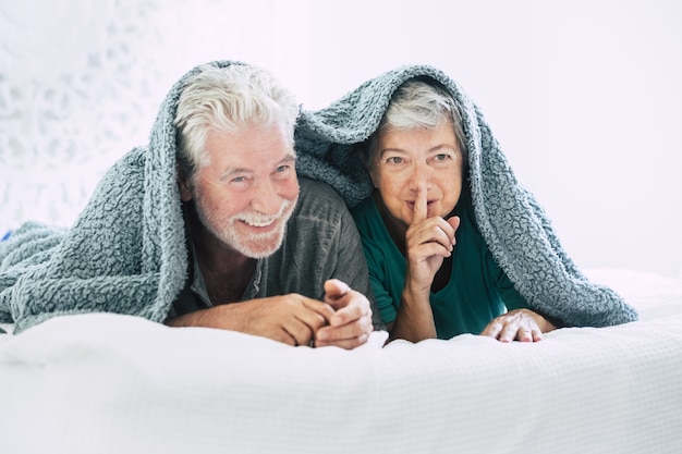 Älteres Paar liegt auf der Vorderseite und gestikuliert mit dem Finger auf den Lippen auf dem Bett. Älteres Ehepaar, das Freizeit miteinander verbringt. Glückliches altes Paar, das auf einem gemütlichen Bett liegt, das mit einer Decke bedeckt ist, mit dem Finger auf den Lippen