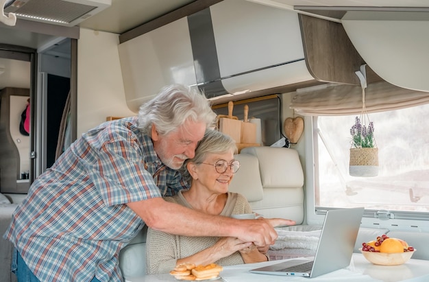 Älteres schönes Paar im Reiseurlaub in einem Wohnmobil, das auf dem Laptop surft, während es gemeinsam frühstückt