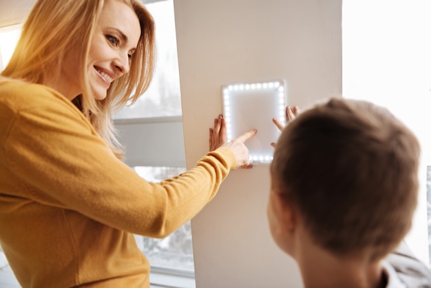 Últimas inovações. Mulher feliz e feliz encantada sorrindo e usando um painel de controle de casa inteligente ao lado de seu filho