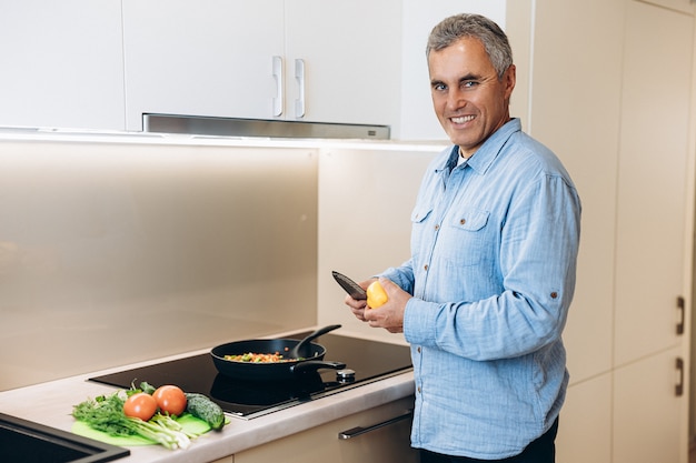 Último homem cozinhando na cozinha. Homem alegre e bonito com cabelo grisalho corta pimentas para adicionar ao seu prato vegetariano. Legumes picados na frigideira. Coma mais verduras e vegetais!