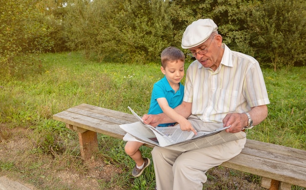 Último homem lendo jornal e linda criança apontando um artigo com o dedo sentado no banco do parque. Conceito de duas gerações diferentes.