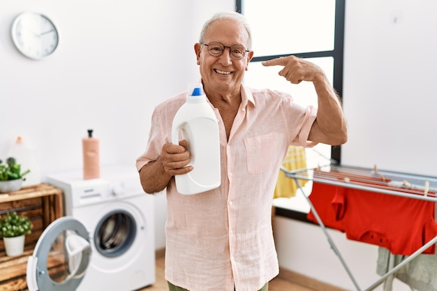 Último homem segurando uma garrafa de detergente na lavanderia apontando o dedo para si mesmo sorrindo feliz e orgulhoso