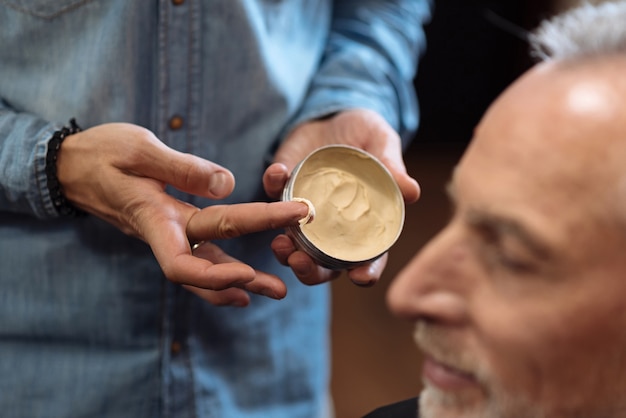 Últimos passos. Perto do cabeleireiro masculino segurando o frasco com gel de cabelo antes de colocá-lo no cliente sênior na barbearia.