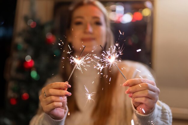 Luces de bengala palo de fuego de bengala en manos femeninas sosteniendo a la mujer en suéter acogedor blanco de lana tejida en el fondo de una casa rodante de camping al aire libre en la noche de año nuevo y nochebuena. chispas chispeantes