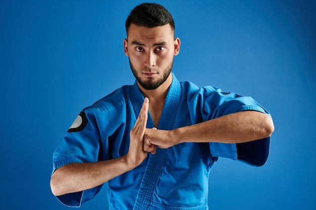 Luchador de karate kazajo asiático en uniforme de kimono que muestra el gesto de las manos sobre fondo azul aislado en estudio