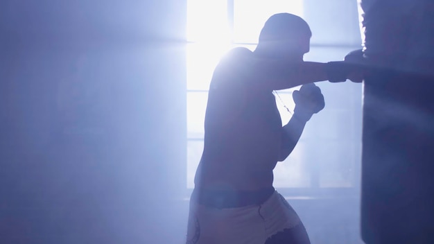 Luchador musculoso y guapo dando una fuerte patada hacia adelante durante una ronda de práctica con una bolsa de boxeo