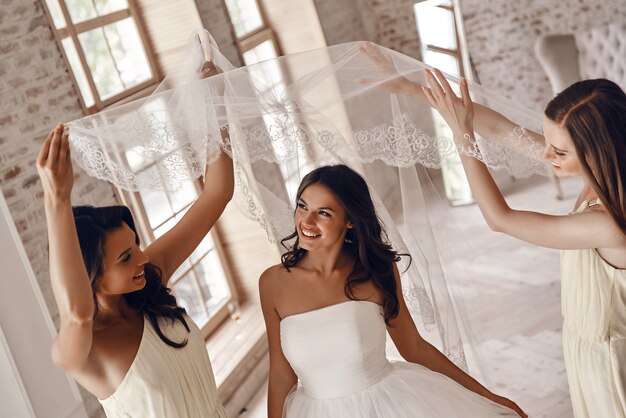 Foto luciendo perfecto. damas de honor ayudando a la novia a ponerse un velo y sonriendo mientras está de pie en el probador