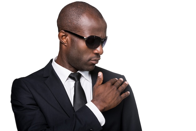 Luciendo simplemente perfecto. Retrato de hombre africano joven serio en ropa formal y gafas de sol ajustando su chaqueta mientras está de pie aislado sobre fondo blanco.