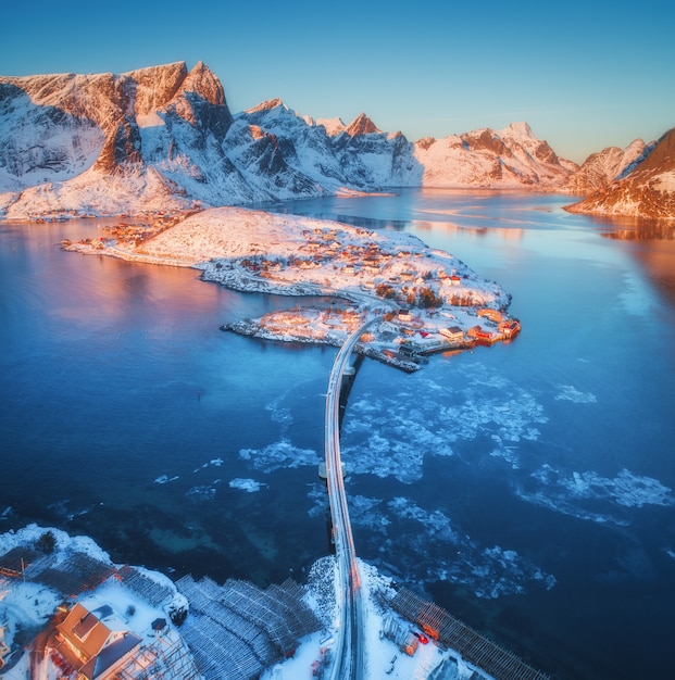 Luftaufnahme der Brücke über dem Meer und den schneebedeckten Bergen in den Lofoten-Inseln, Norwegen. Reine und Hamnoy bei Sonnenaufgang im Winter.