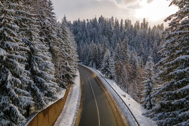 Luftaufnahme der Winterlandschaft mit schneebedeckten Berghügeln und kurvenreicher Forststraße am Morgen.