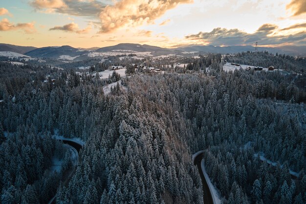 Luftaufnahme der Winterlandschaft mit schneebedeckten Berghügeln und kurvenreicher Forststraße am Morgen.