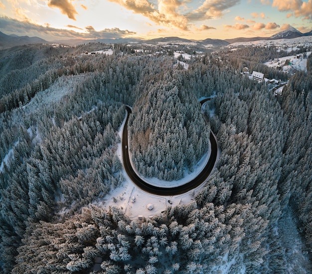 Luftaufnahme der Winterlandschaft mit schneebedeckten Berghügeln und kurvenreicher Forststraße am Morgen.