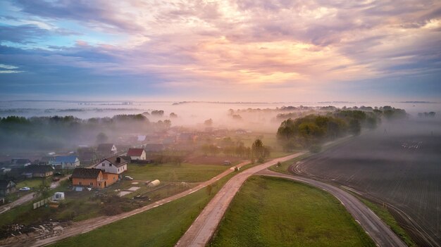 Luftaufnahme des Dorfes, der ländlichen Schotterstraße und der von Nebel bedeckten Bäume. Sonnenaufgangspanorama am frühen nebligen Morgen. Frühling Sommerfelder. Regnerisches trübes Wetter. Weißrussland, Gebiet Minsk