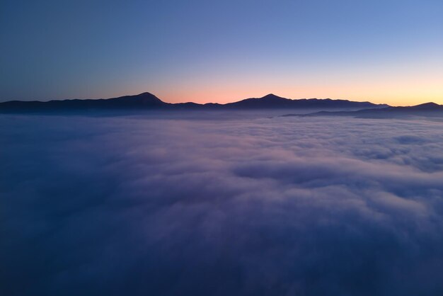 Luftaufnahme des farbenfrohen Sonnenaufgangs über weißem, dichtem Nebel mit entfernten dunklen Silhouetten von Berghügeln am Horizont