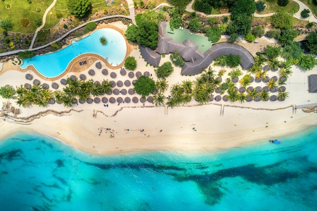 Luftaufnahme des tropischen Sandstrandes mit Palmen und Sonnenschirmen an sonnigen Tagen Sommerurlaub am Indischen Ozean Sansibar Afrika Landschaft mit Palmen Hotels Pool weißer Sand azurblaues Meer Draufsicht