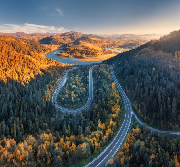 Luftaufnahme einer Bergstraße in einem farbenfrohen Wald bei Sonnenuntergang im Herbst in der Ukraine. Draufsicht von der Drohne aus auf eine Straße in einem Wald im Herbst. Wunderschöne Landschaft mit Autobahn in Hügeln, Flusskiefern, Bergen