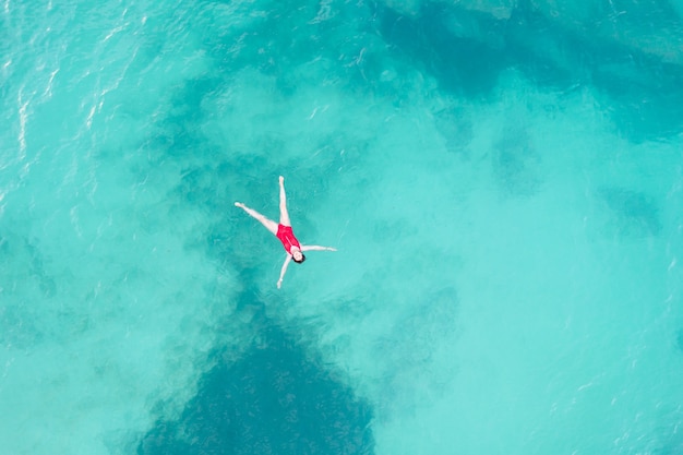 Luftaufnahme einer Frau mit rotem Badeanzug, die auf einer transparenten türkisfarbenen Wasseroberfläche liegt