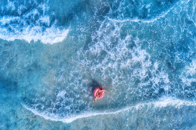 Luftaufnahme einer jungen Frau, die an einem sonnigen Sommertag mit dem rosa Donut-Schwimmring im klaren blauen Meer mit wunderschönen Wellen schwimmt. Tropische Luftlandschaft mit azurblauem Mädchenwasser. Draufsicht auf Reisen