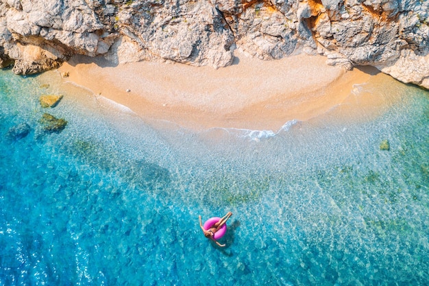 Luftaufnahme einer jungen Frau, die mit einem rosa Donut-Schwimmring im klaren blauen Meer mit Wellen, Sandstrand und Felsen bei Sonnenuntergang im Sommer schwimmt. Tropische Landschaft mit azurblauen Wassersteinen für Mädchen. Draufsicht